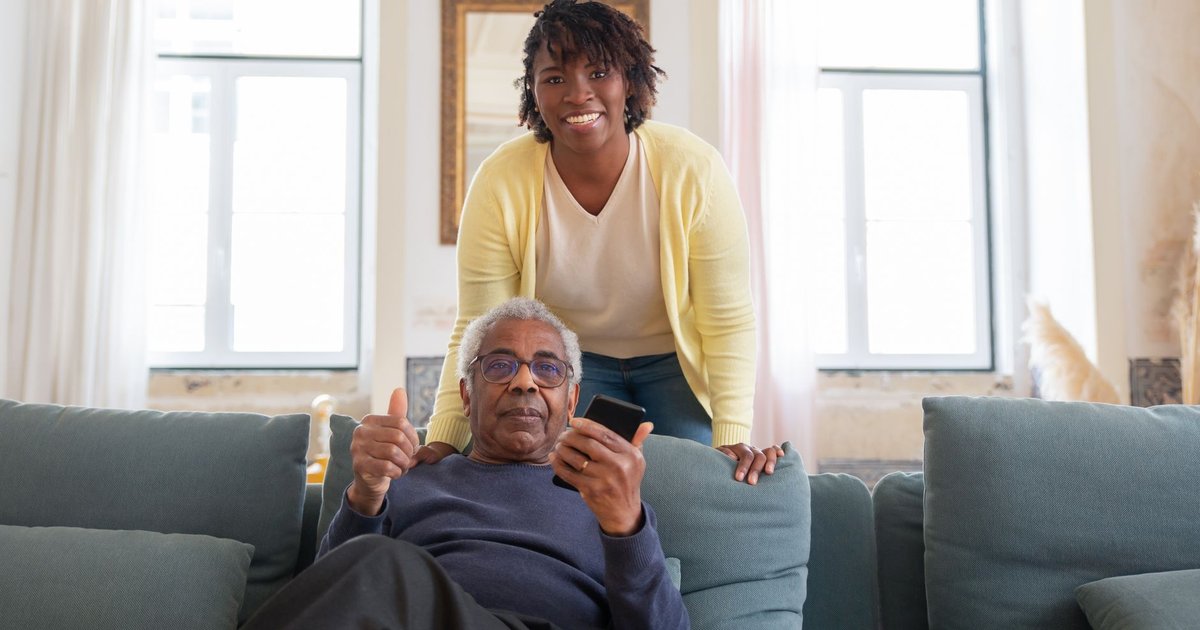 Smiling woman in a yellow cardigan stands behind an older man holding a phone