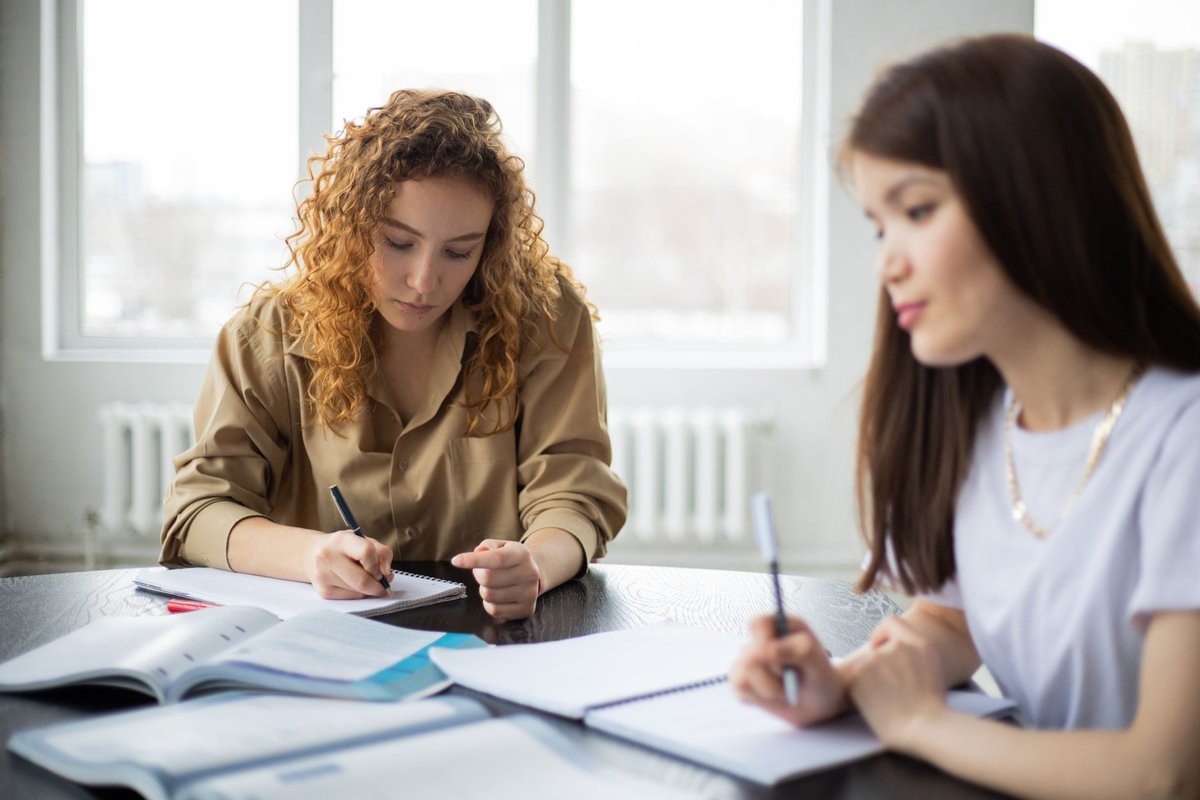 Two women taking notes at a table with textbooks in a bright room