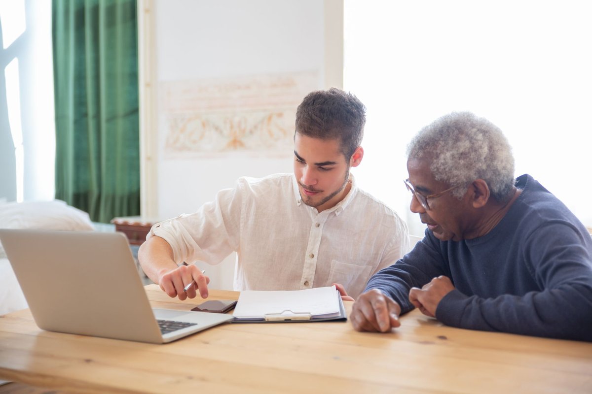 Young man pointing at a clipboard beside a laptop while an older man looks on