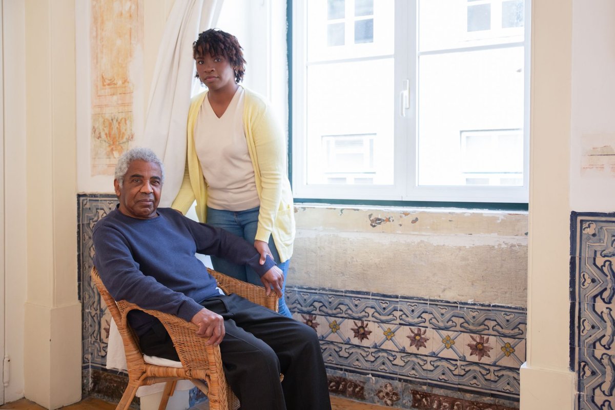 Caregiver standing beside an older man seated in a wicker chair by a window