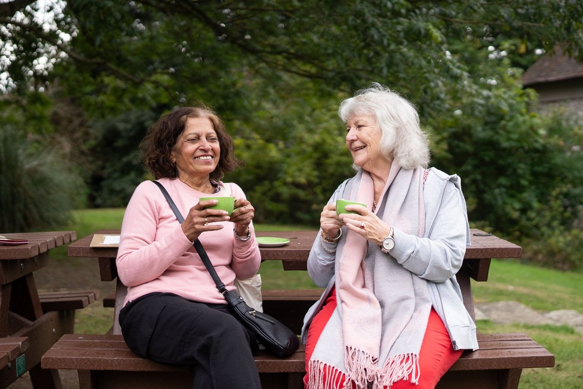 Two older women laughing together over cups of tea at a park picnic table
