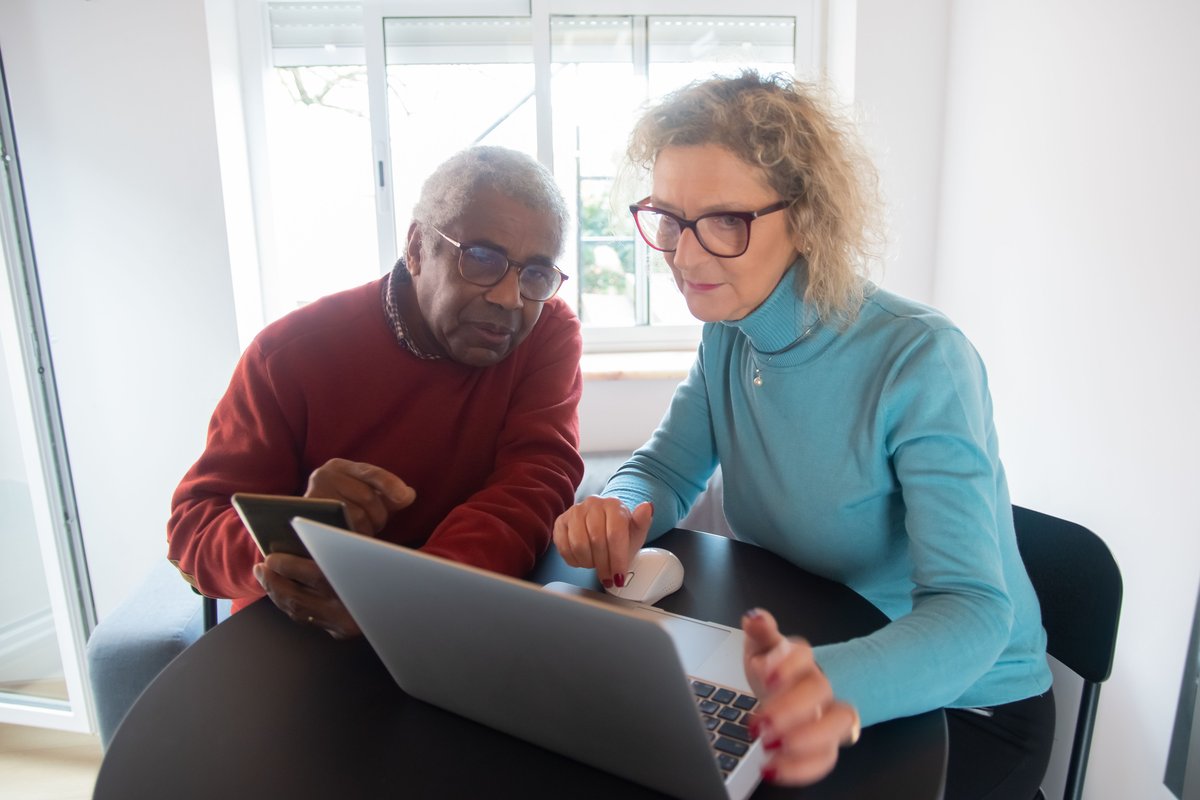 Older man holding a phone beside a woman using a laptop at a round table
