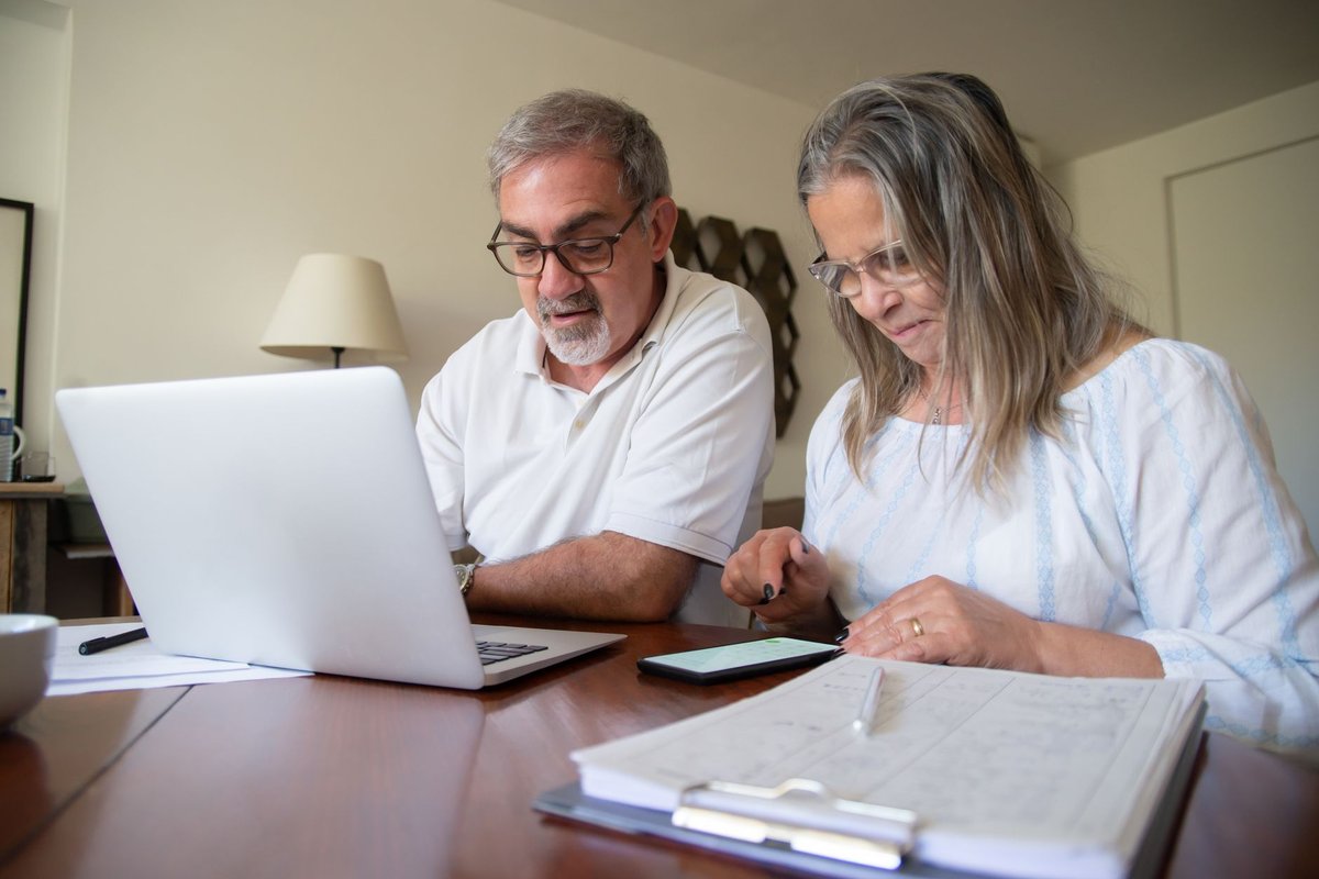 Older couple sitting at a wooden table with a laptop, phone and paperwork
