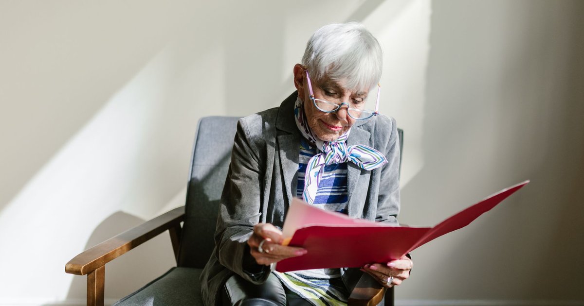 Older woman with white hair reading papers from a red folder in an armchair