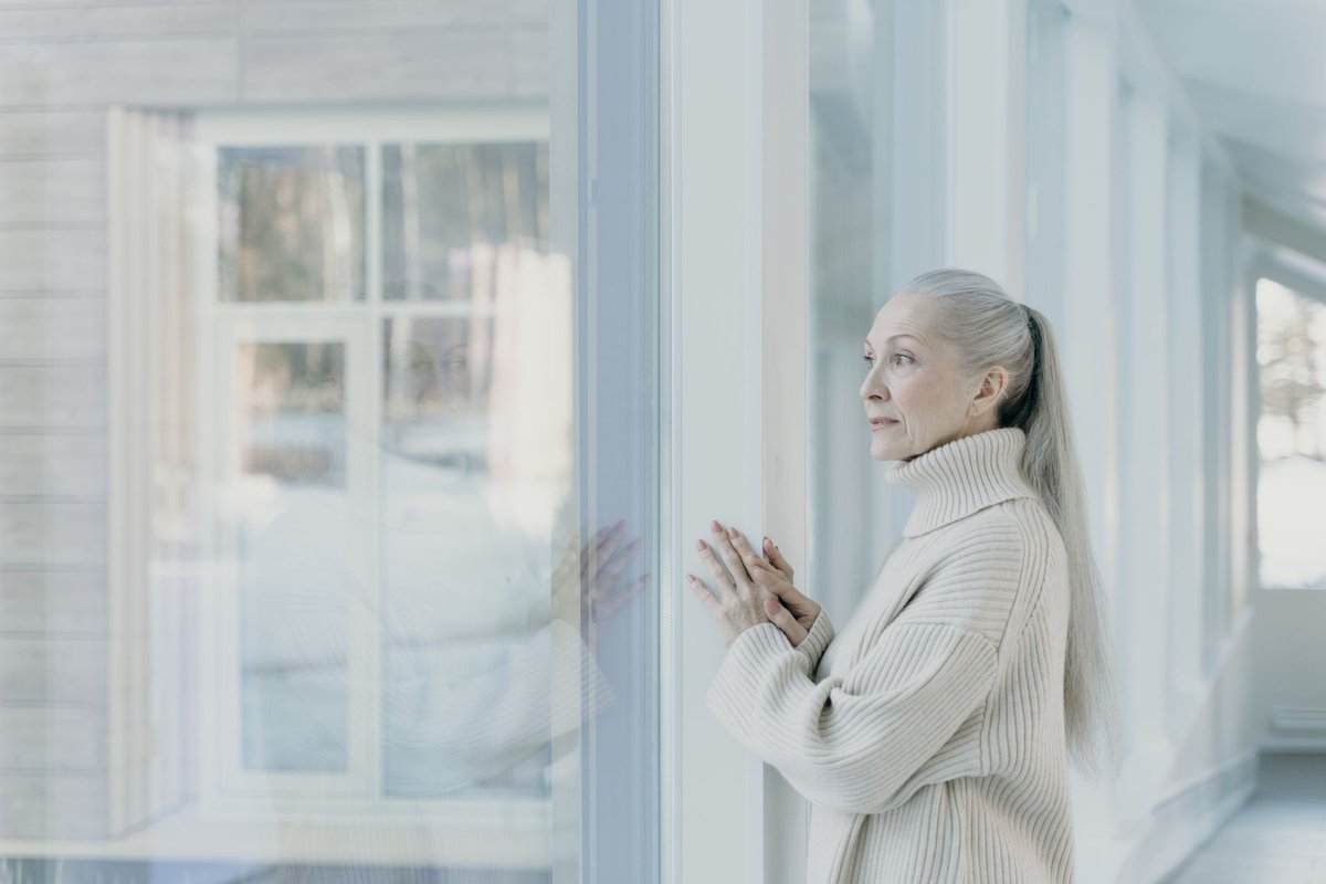 Older woman standing at a bright window, looking out alone on a quiet winter day