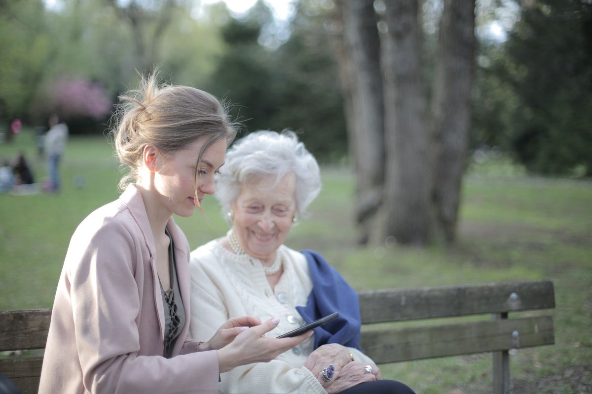 Young woman shows an older woman a smartphone while they sit together on a park bench