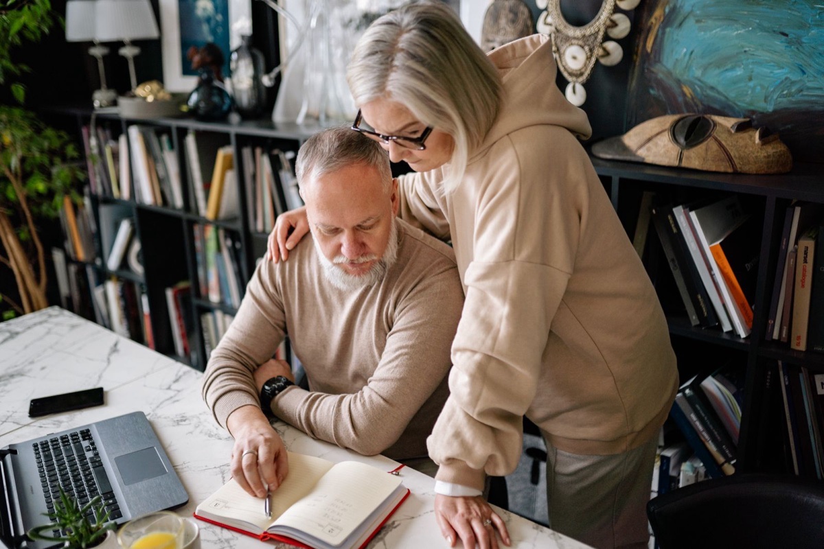 Older couple writing in a notebook together at a table with a laptop