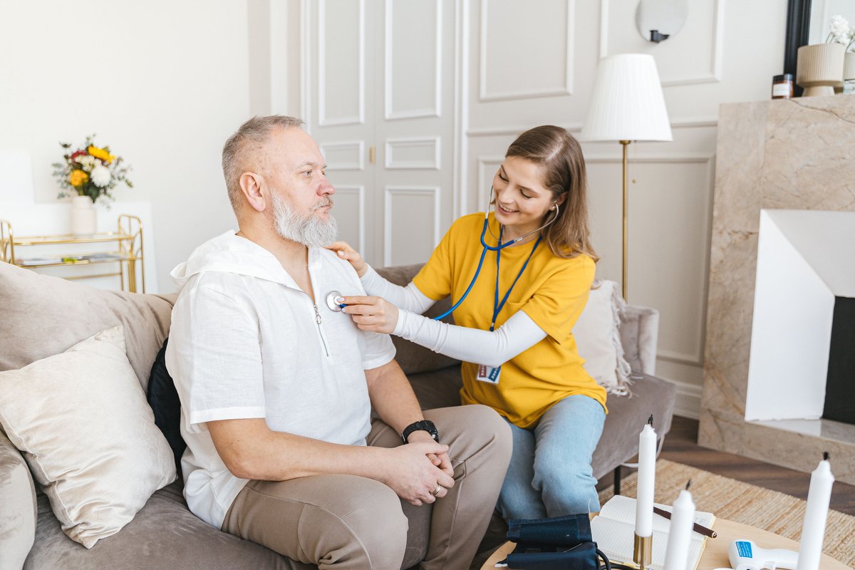 Nurse with a stethoscope doing a home check-up on an older man on a sofa
