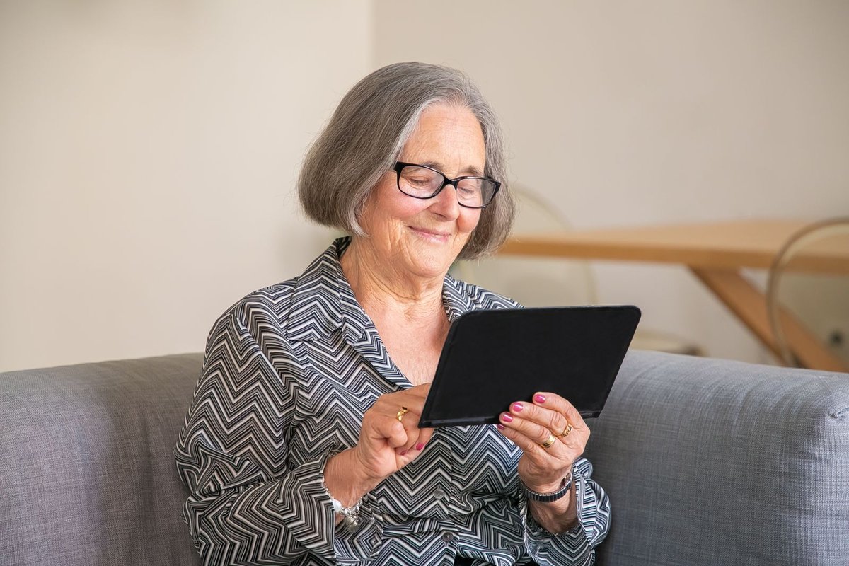 Smiling older woman with glasses using a tablet while sitting on a gray sofa at home