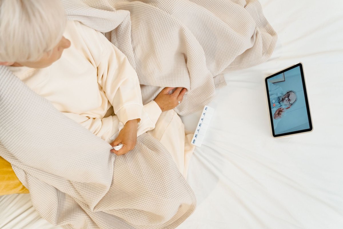 Older woman in bed talking to a doctor on a tablet video call, pill organizer beside her