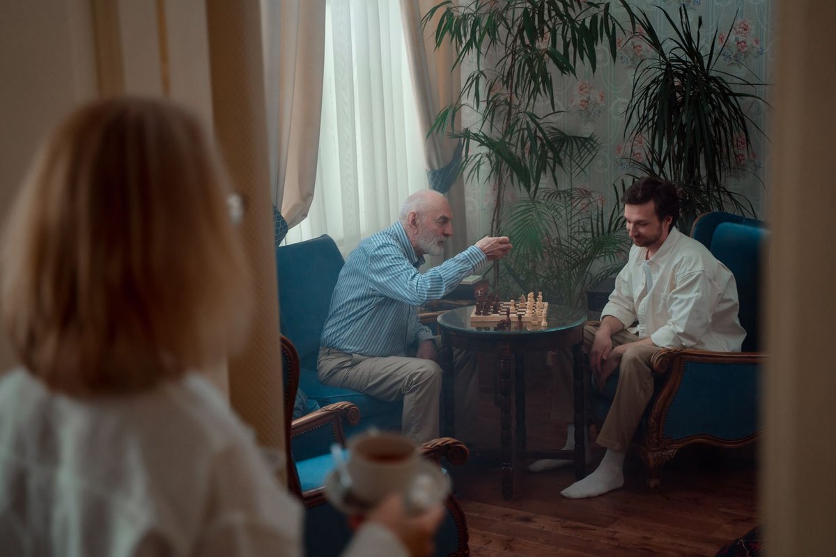 Older man and an adult son playing chess in a living room while a woman with a teacup watches