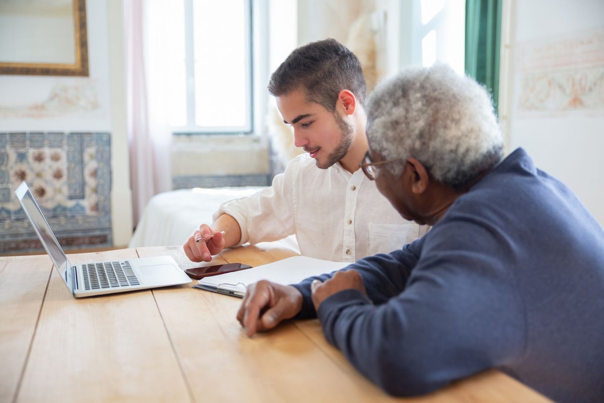 Young man helping an older man with a laptop and papers at a wooden table