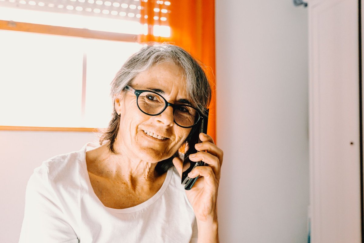 Smiling older woman with glasses talking on a mobile phone at home by a bright window