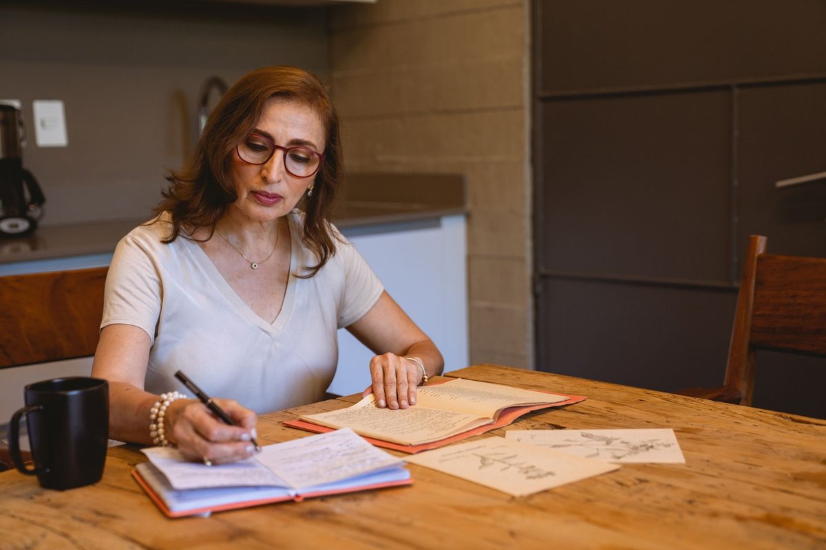 Woman with glasses writing in a notebook at a wooden kitchen table beside a mug