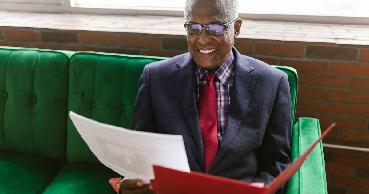 Smiling older man in glasses reading papers from a red folder on a green sofa