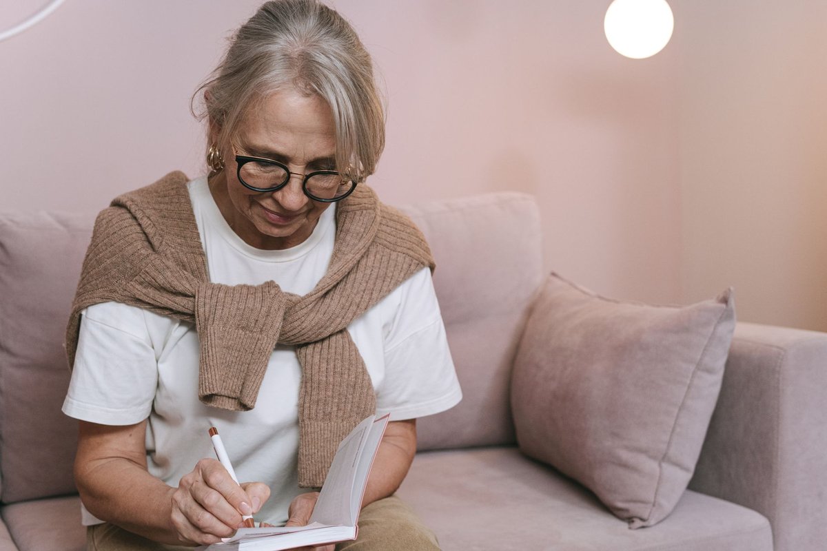 Older woman with glasses writing a list in a small notebook while sitting on a sofa