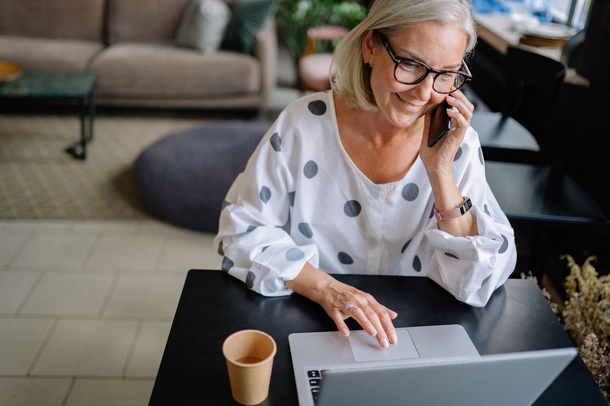 Smiling woman with glasses talking on her phone while using a laptop at a table