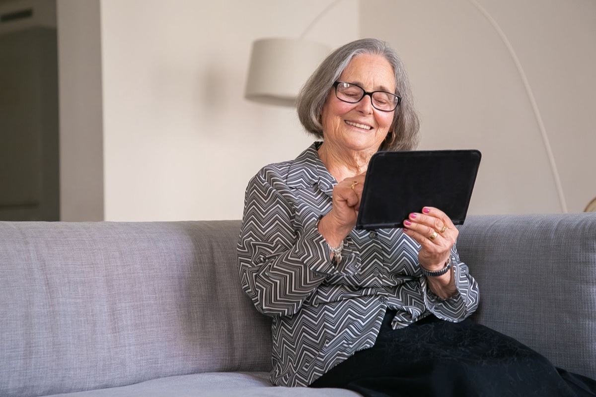 Smiling older woman with glasses using a tablet on a gray sofa at home