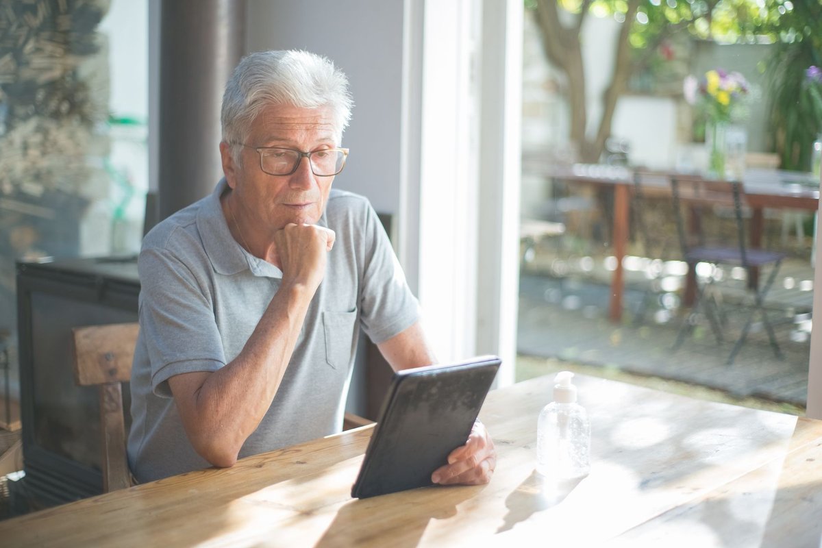 Gray-haired man in glasses reading a tablet at a sunny wooden table by a window