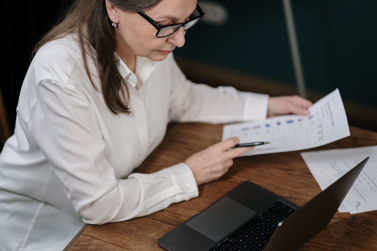 Woman with glasses reviewing papers with a pen next to an open laptop
