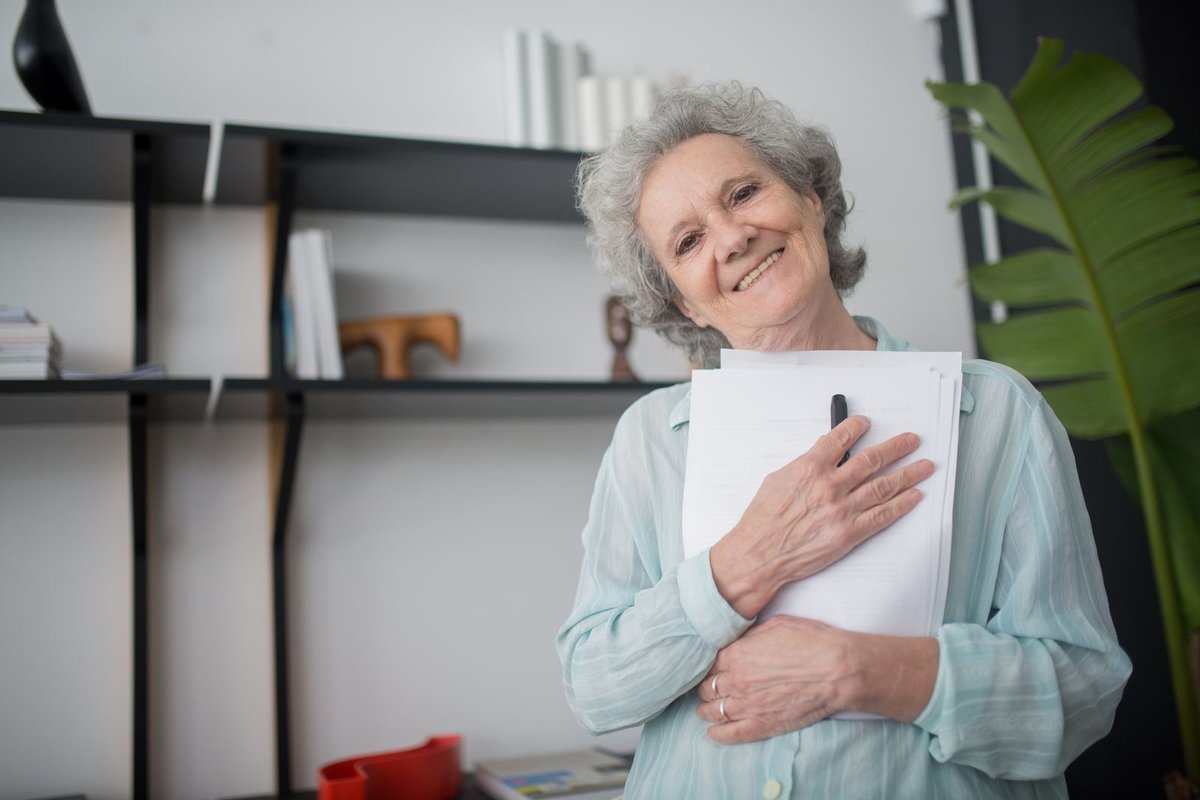 Smiling gray-haired woman holding a stack of papers and a pen in a bright room