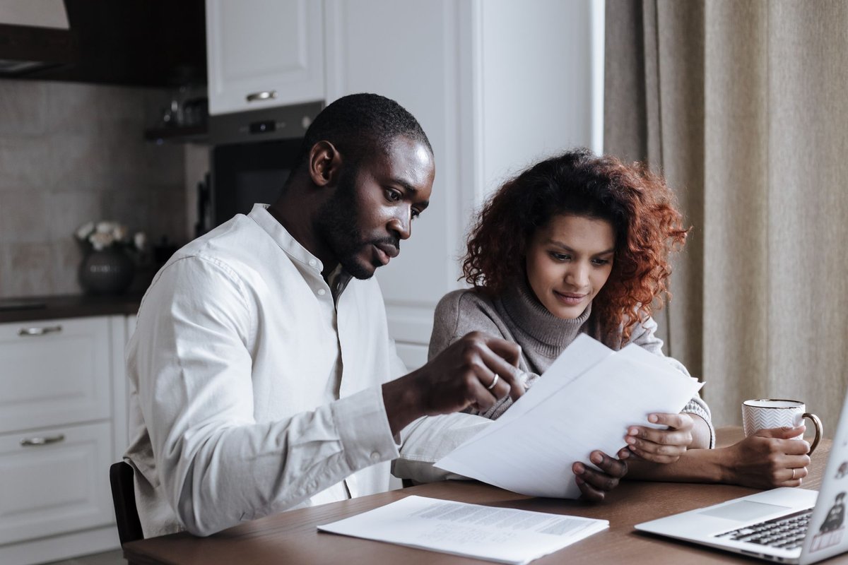 Two adults reading paperwork together at a kitchen table with a laptop and coffee
