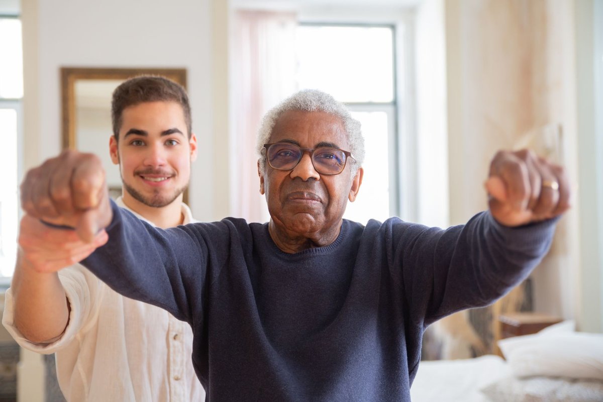 Older man doing an arm exercise at home while a younger helper stands behind him