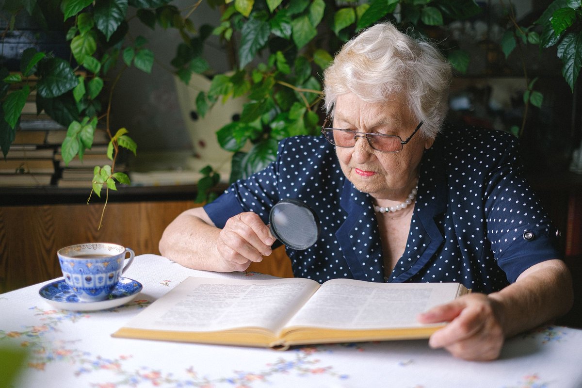 Older woman in glasses using a magnifying glass to read a book at a table