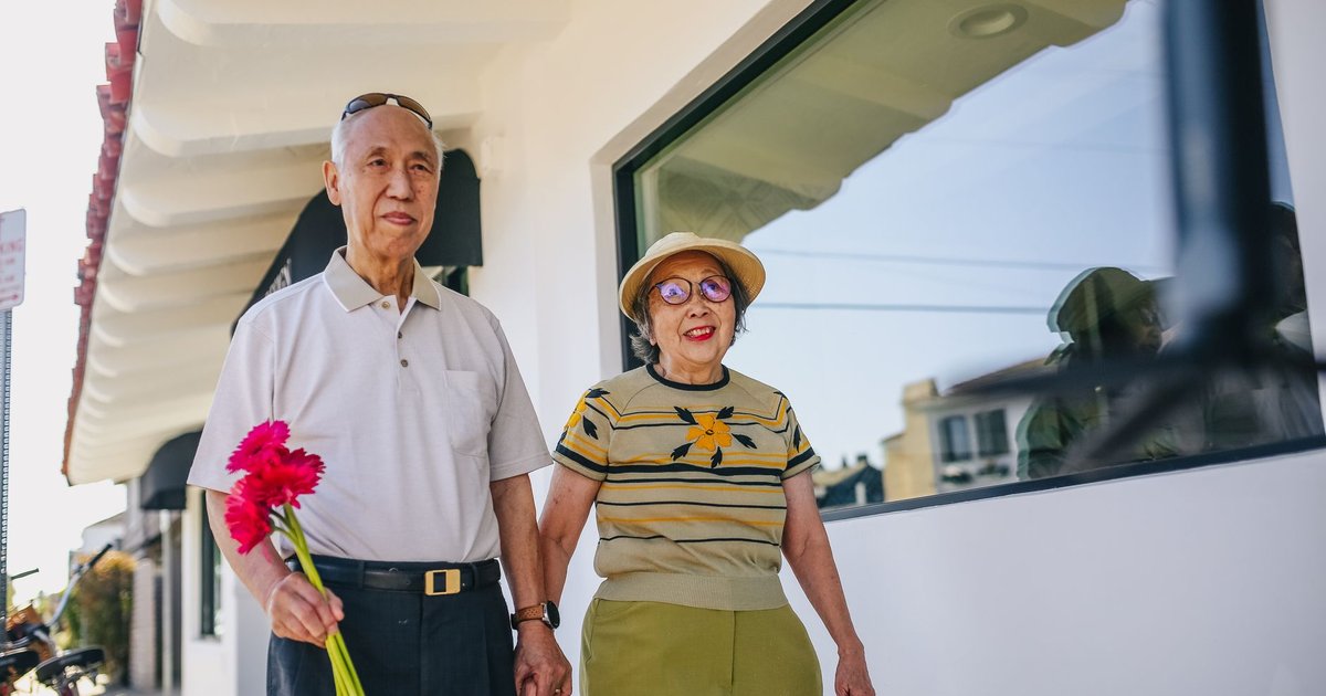 Older couple walking hand in hand down a sunny street, he holds pink flowers