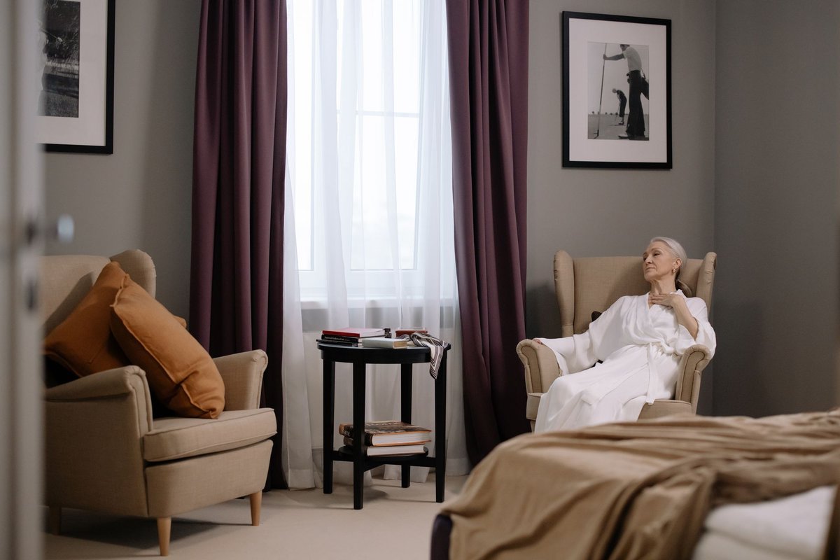 Older woman resting in a cream armchair beside a window in a bright bedroom