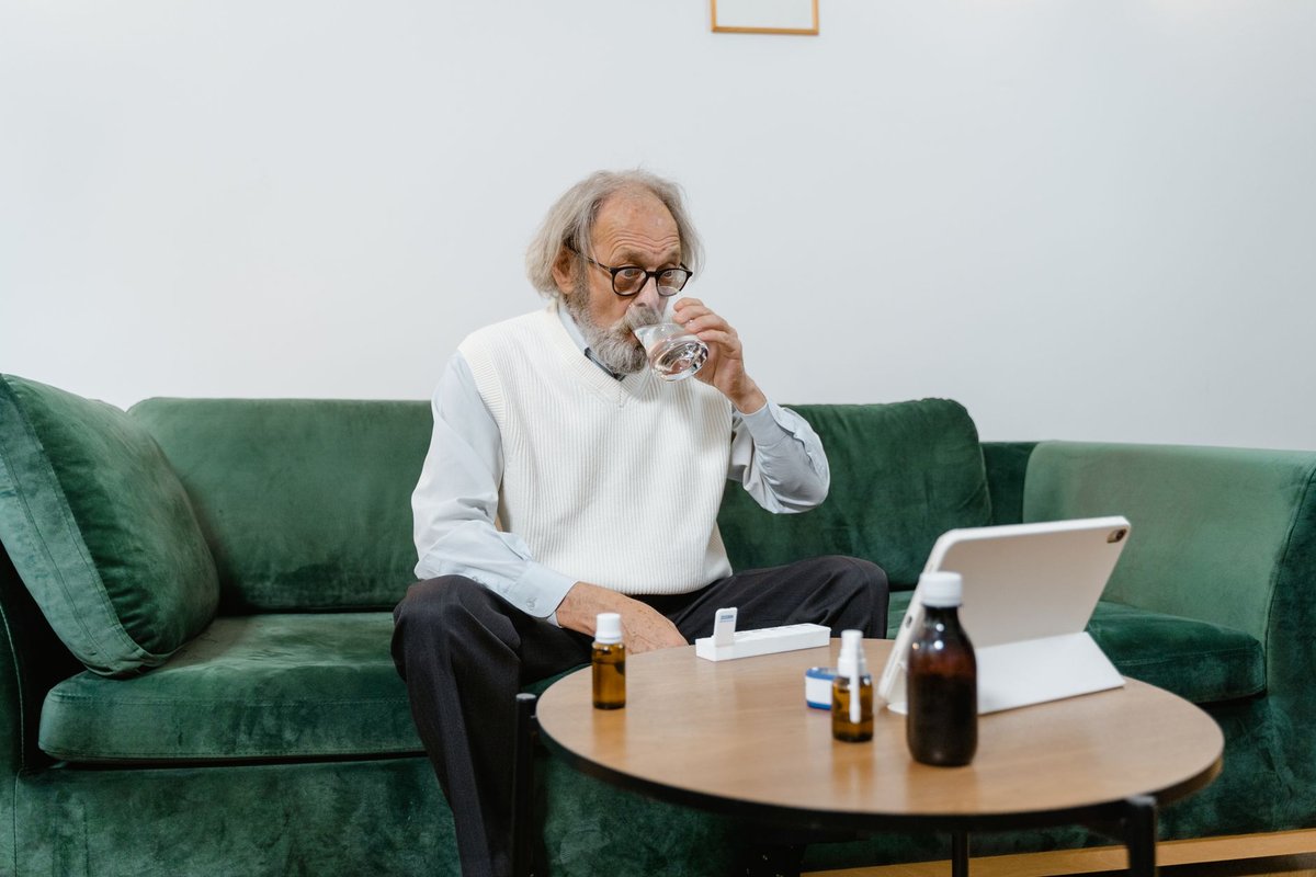 Older man at home drinking water with medicine bottles and a pill box on the table