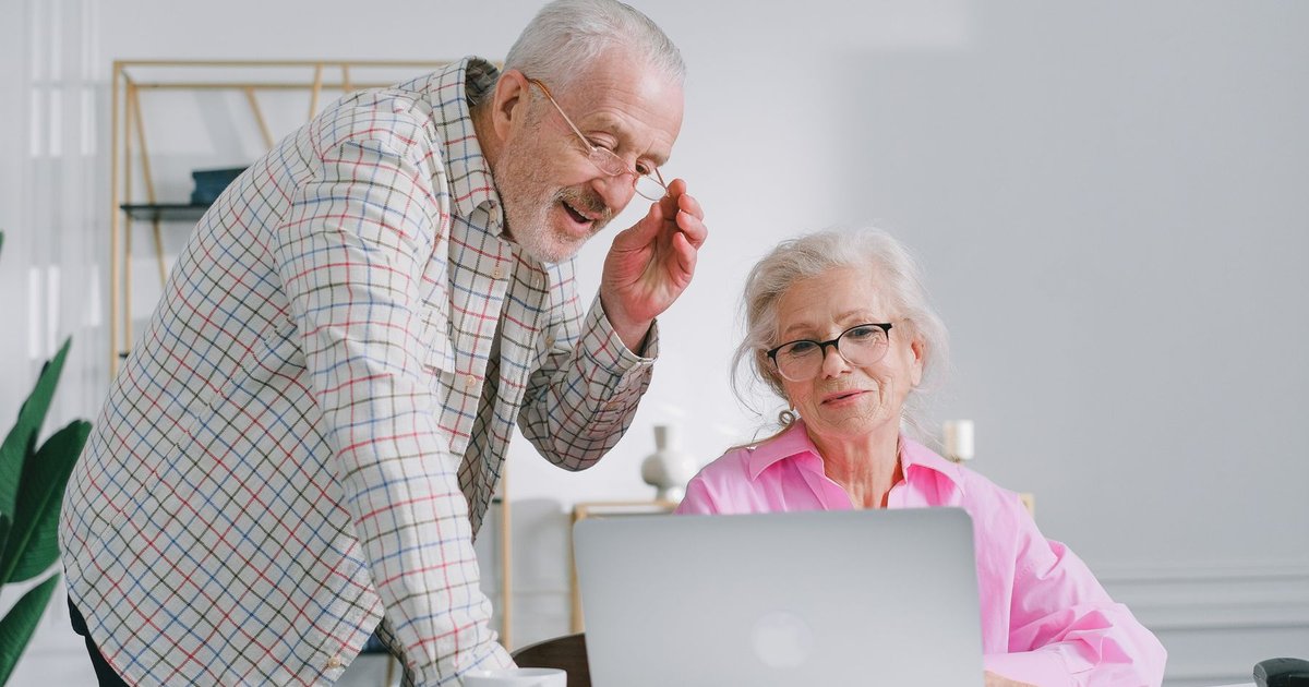 Older man in glasses leaning over to look at a laptop with his wife at a table
