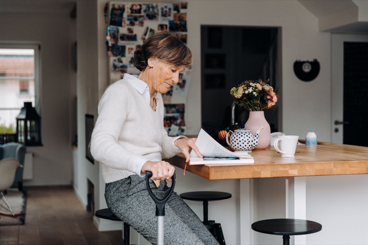 Older woman in a bright kitchen leaning on a stool and paging through a book