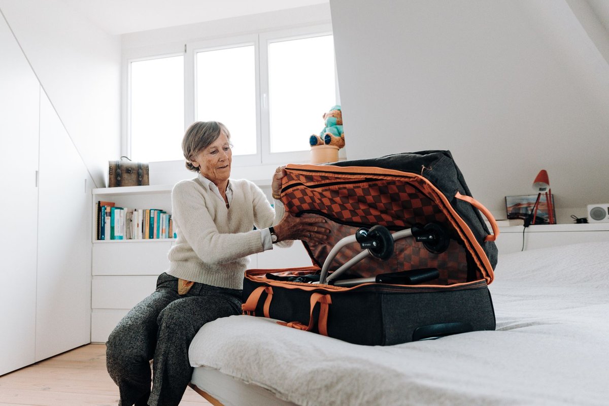 Older woman sitting on a bed in a bright bedroom unpacking a suitcase