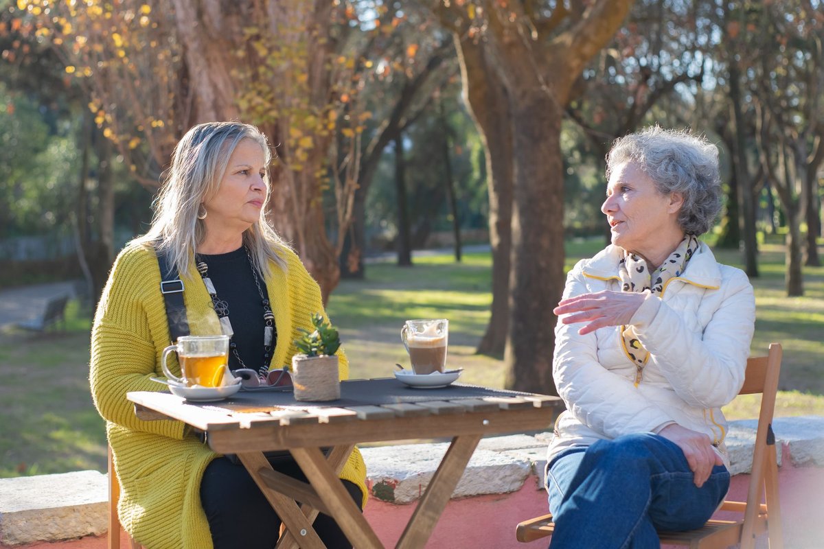 Two older women talk at a small outdoor table with cups of tea