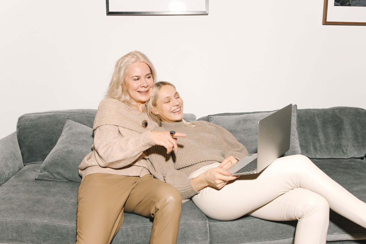 Mother and adult daughter smiling at a laptop together on a gray sofa