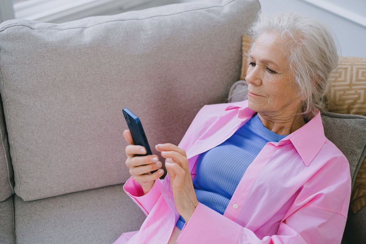 Older woman reading something on her smartphone while seated on a gray sofa