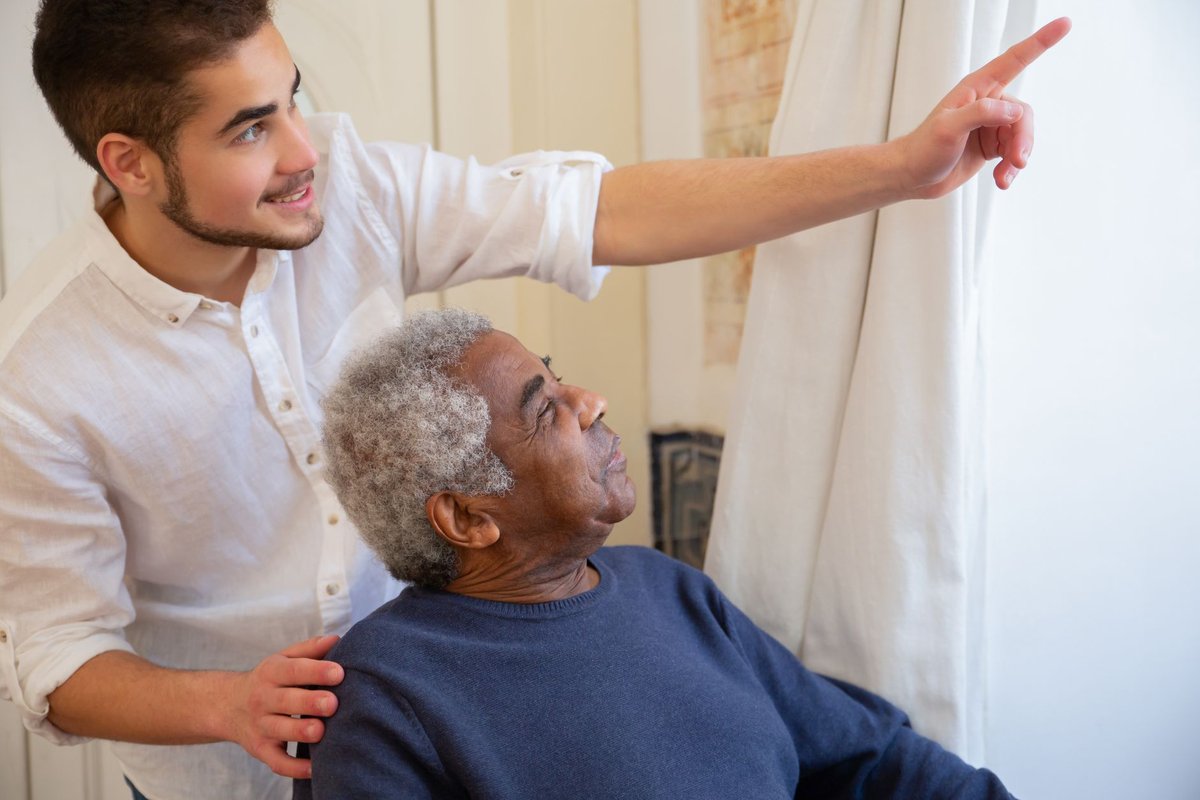 Young caregiver and an older man looking out a bright window together at home