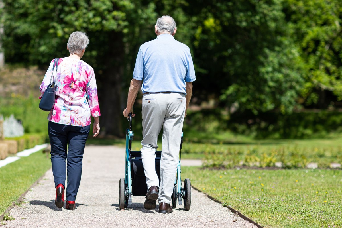 Older couple walking along a gravel park path, one using a wheeled walker