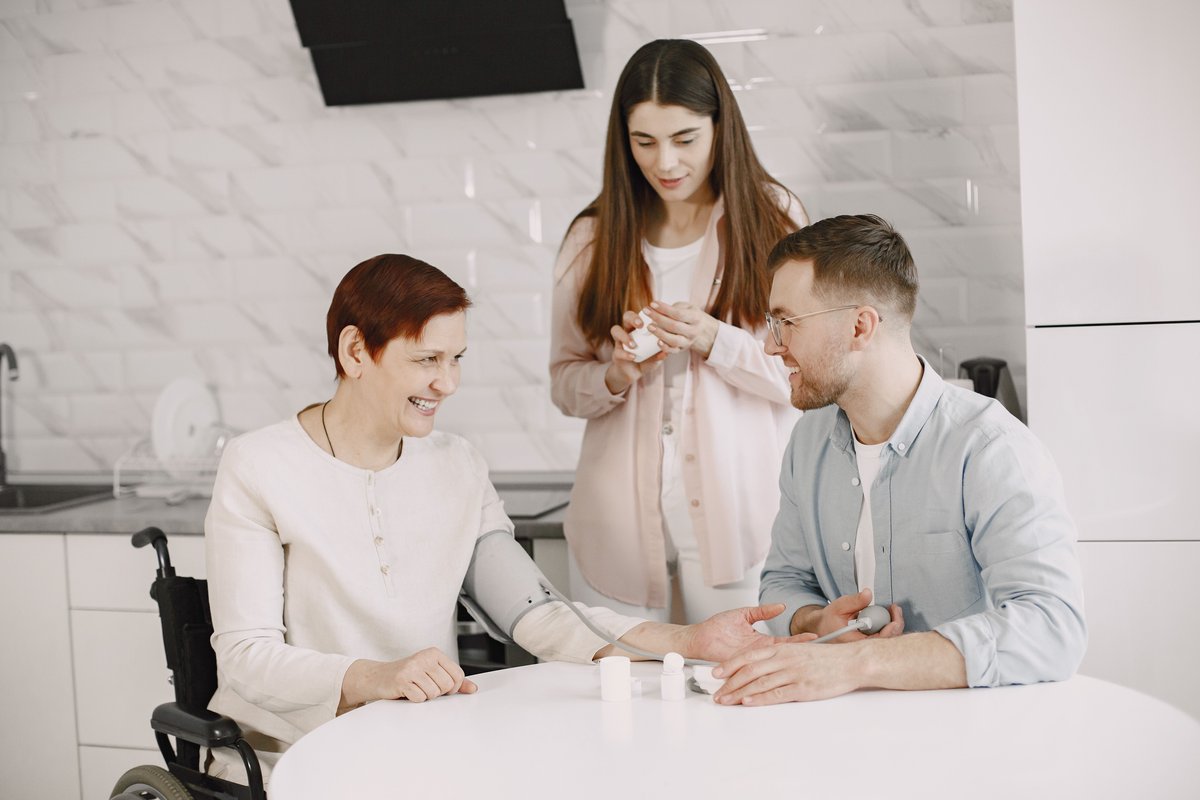 Woman in a wheelchair smiling while a man checks her blood pressure in a kitchen