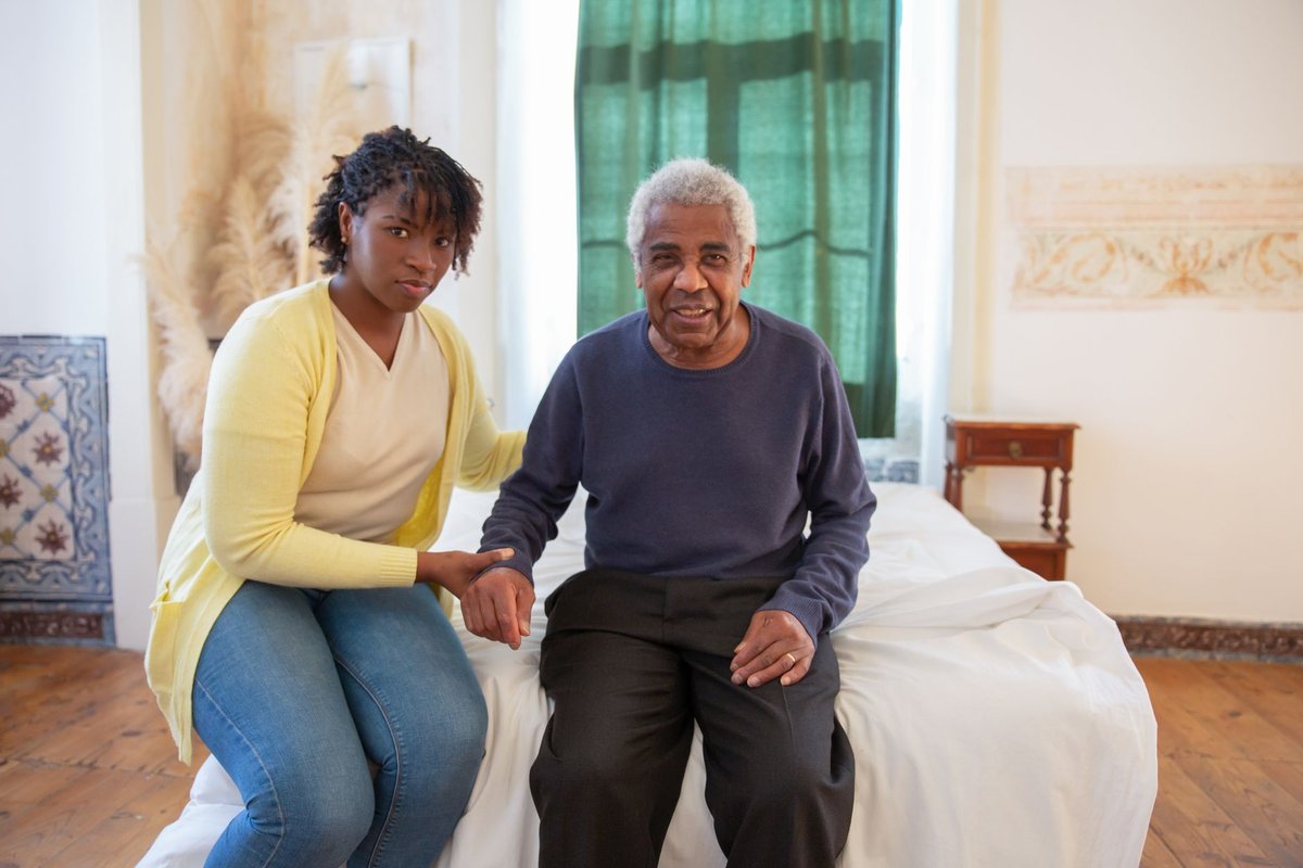 Home aide sitting beside an older man on the edge of his bed, holding his hand
