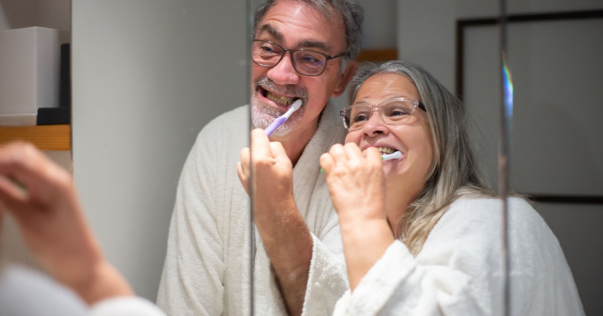 Older couple smiling while brushing their teeth together at a bathroom mirror