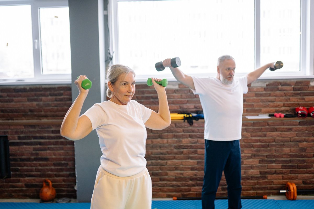 Older man and woman lifting small dumbbells together in a bright exercise room