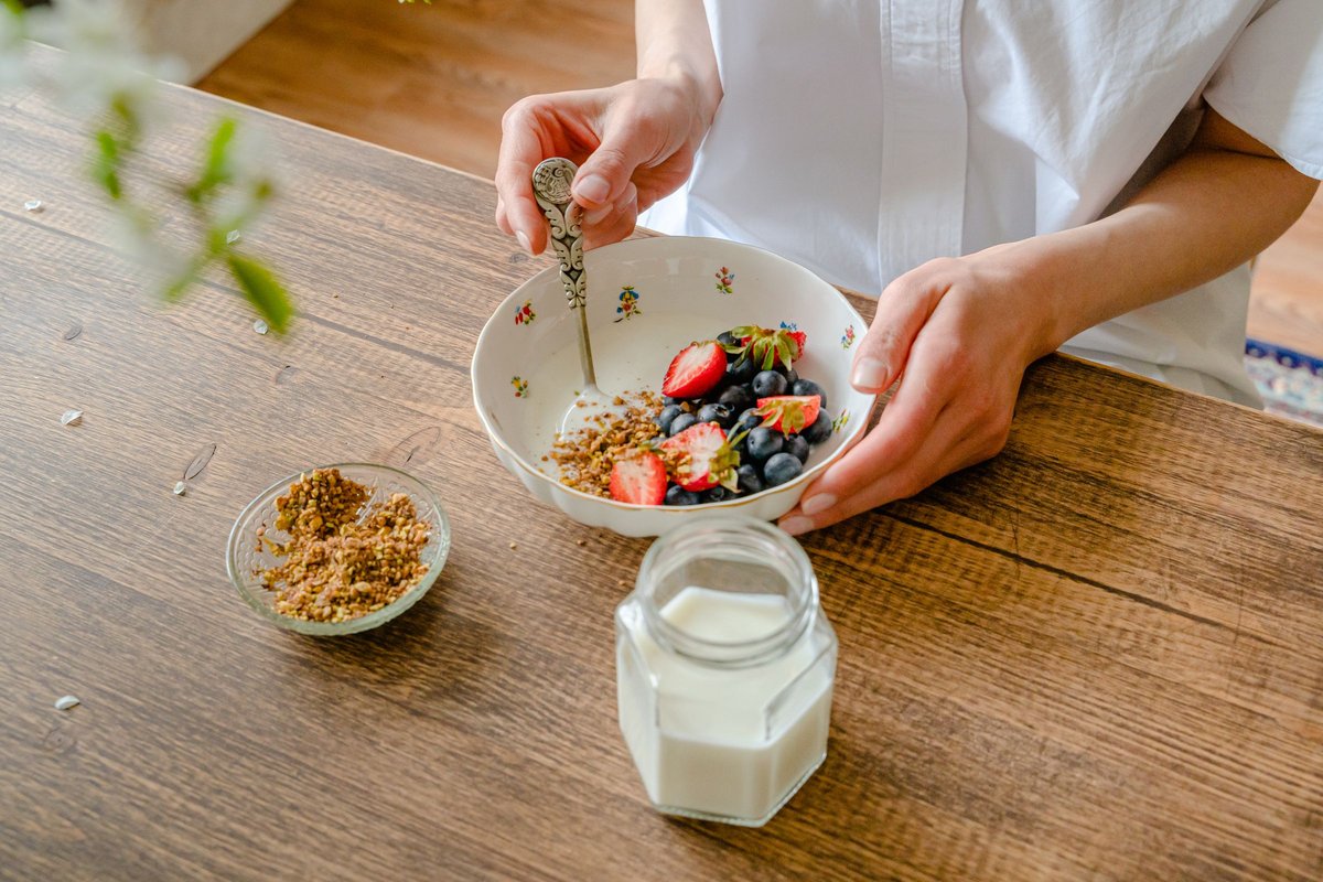 Hands spooning berries and granola into a bowl of yogurt beside a jar of milk