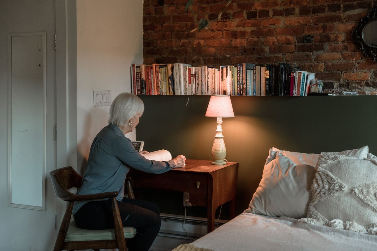 Older woman sitting at a small desk beside a bed in the warm light of a table lamp