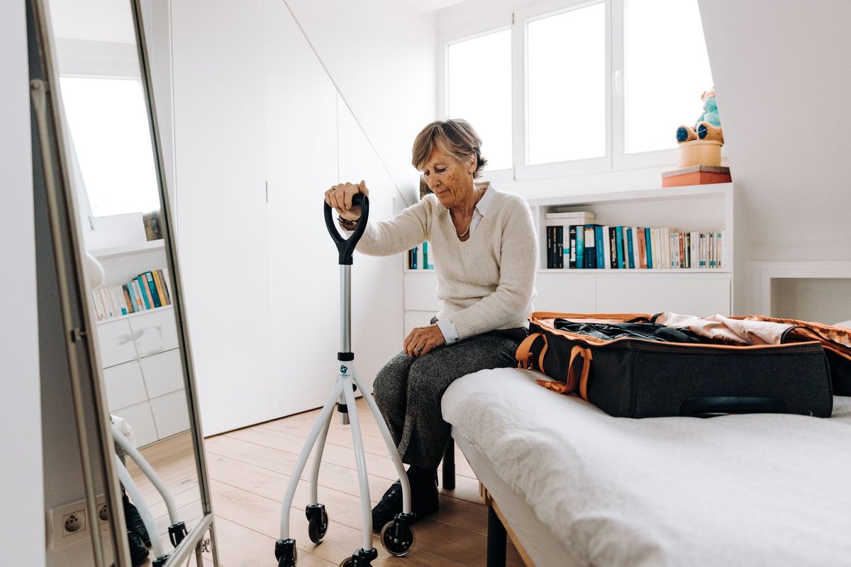 Older woman sitting on the edge of a bed holding a four-footed cane in a bright bedroom
