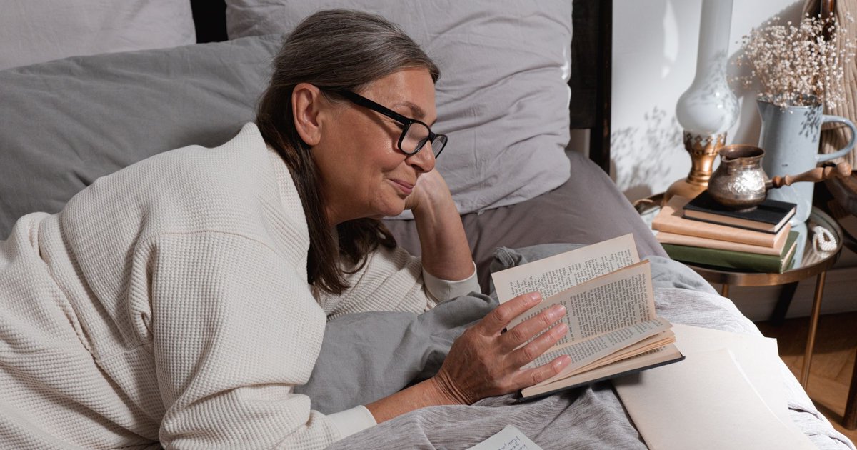 Older woman with glasses reading a book in bed, wrapped in a cozy cream sweater