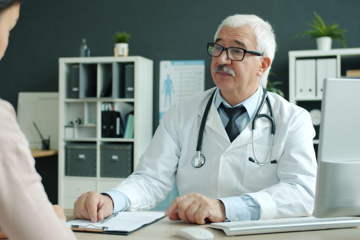 Senior doctor in a white coat talking with a patient across a desk in a bright office