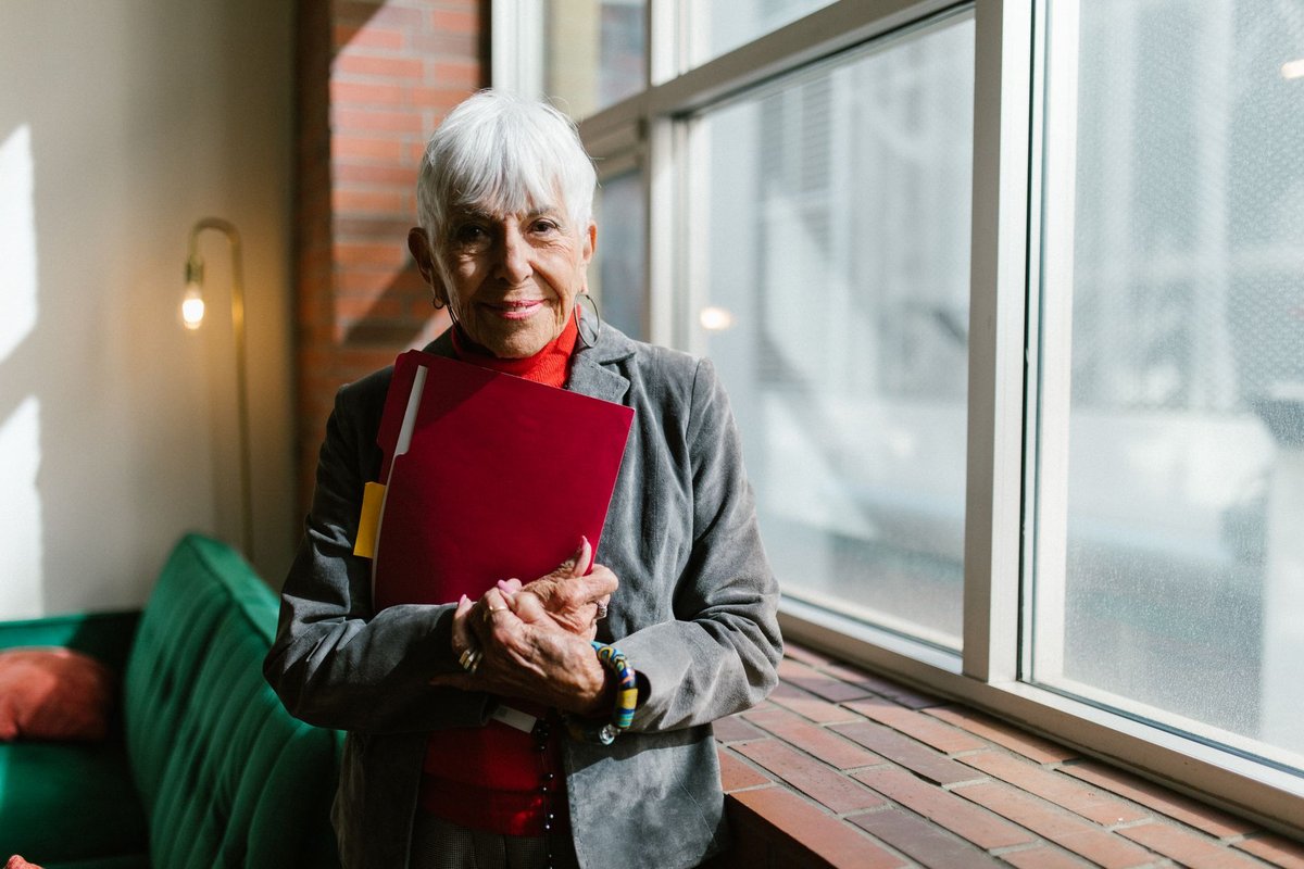 Smiling older woman holding a red folder of papers beside a bright window