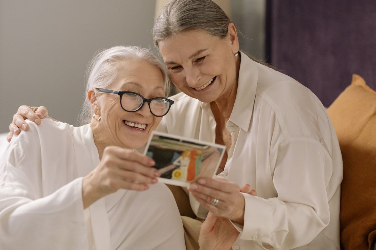 Two smiling older women looking at a printed photo together on a sofa